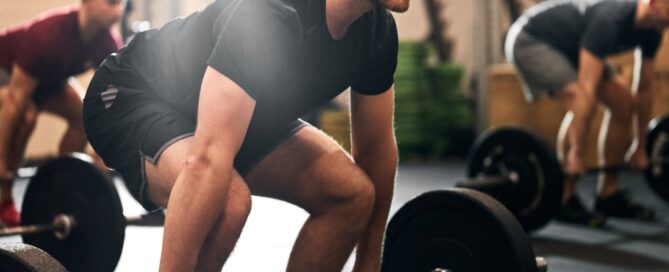 There is a man about to lift up a bar with weights on both sides. He is wearing black and grey clothes and there are two individuals behind him also about to lift the weight bar. They are in a gym.