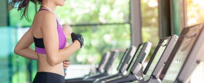 There is a woman in a purple and black sports bra and black pants running on a treadmill. There is a line of them so she is in the gym and she has a black glove on one hand and has white ear buds in.