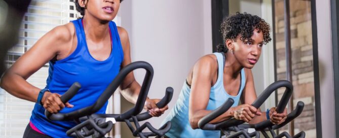 There are two African American women on stationary bikes with blue tops. They have determined expressions on their faces and have light grey walls behind them.