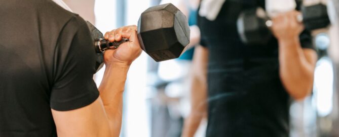There is a man holding a weight up. The man is wearing a black shirt and shorts and has a white towel on his shoulders. He is looking in a mirror and you can see his reflection and the reflection of a gym with people working out.